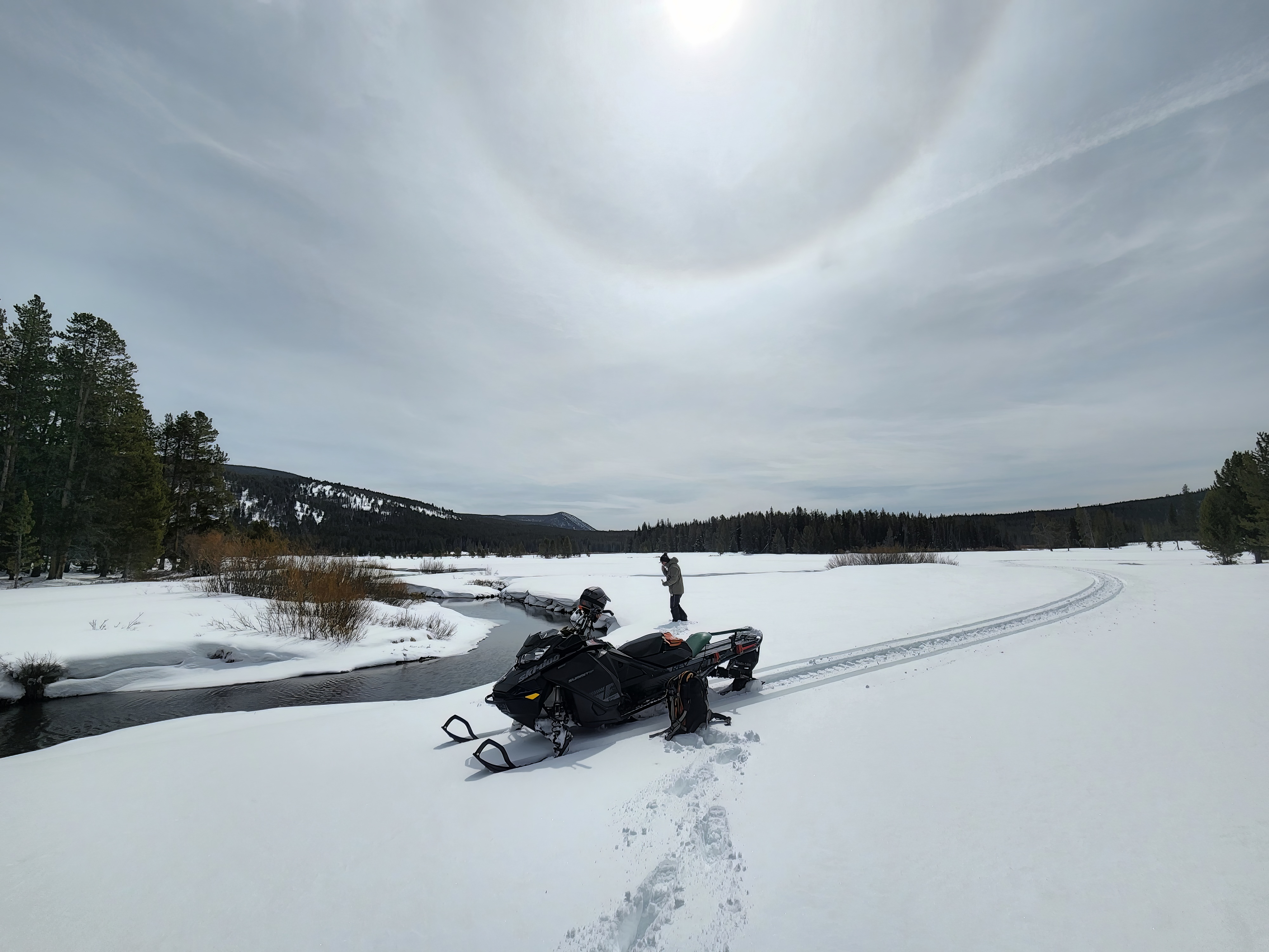 A snowy landscape with a snow mobile parked by a river where someone is walking to collect a water sample.