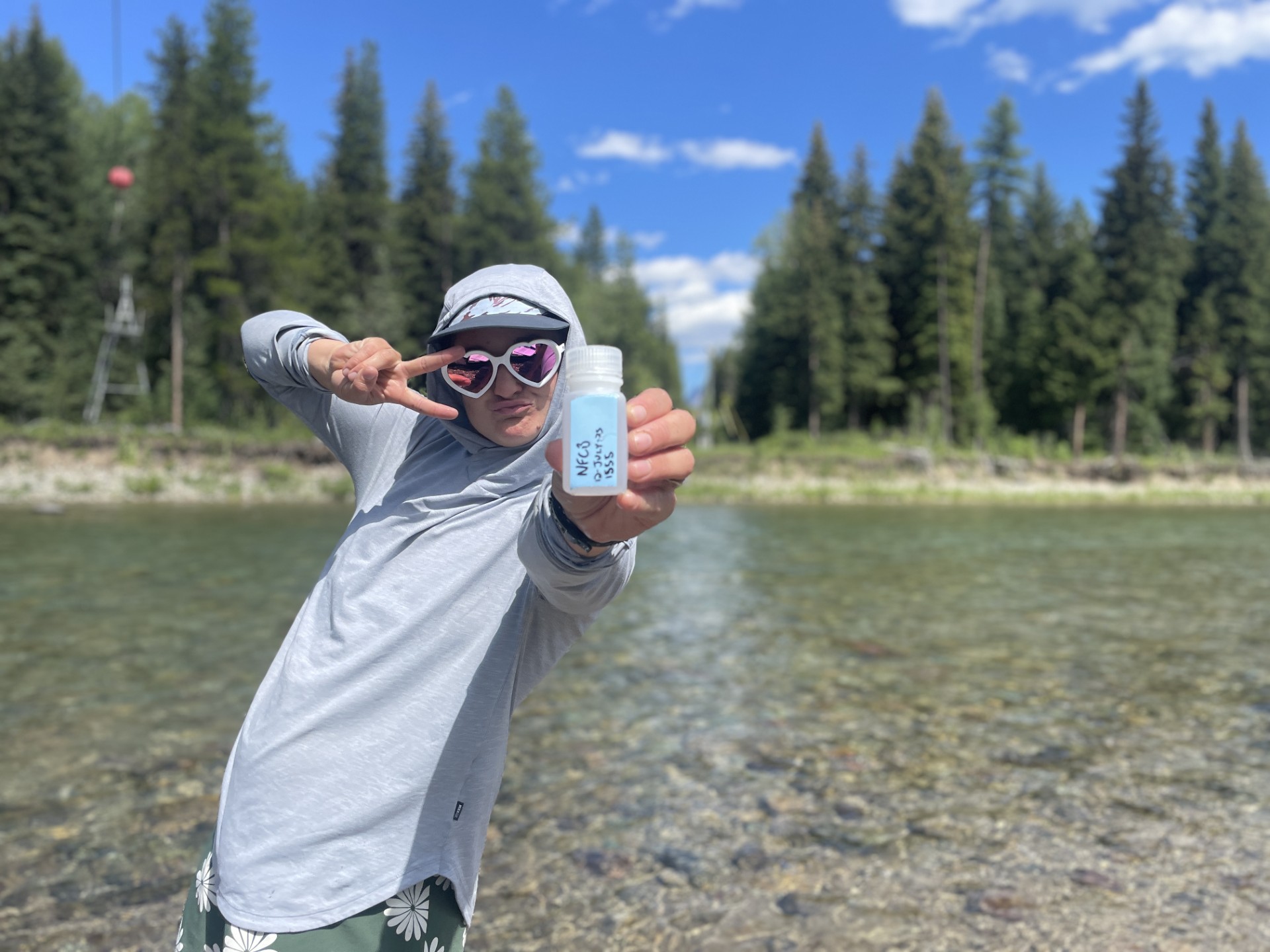 Flathead Rivers Alliance member posing with a “grab” water sample in front of a river and tree line.