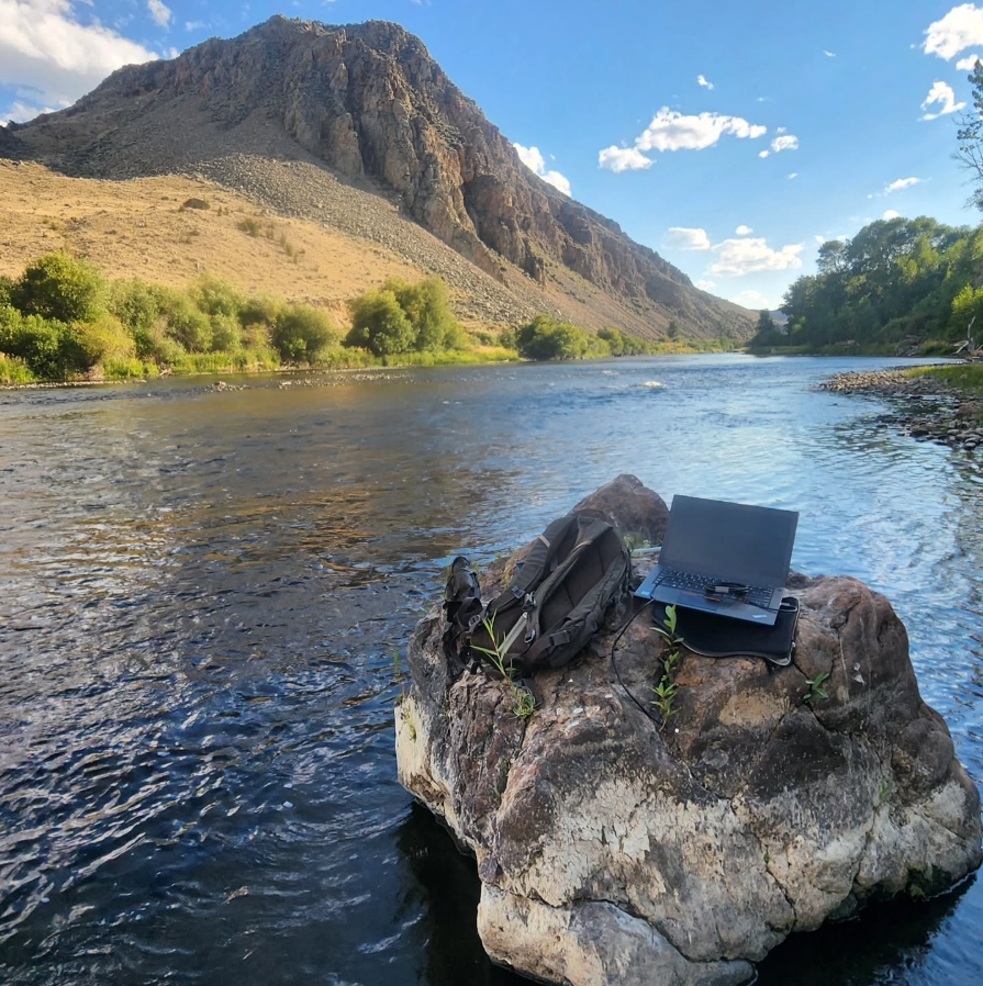 A computer and backpack sitting on a rock in a river with a mountain range in the background where Save Wild Trout collects samples.