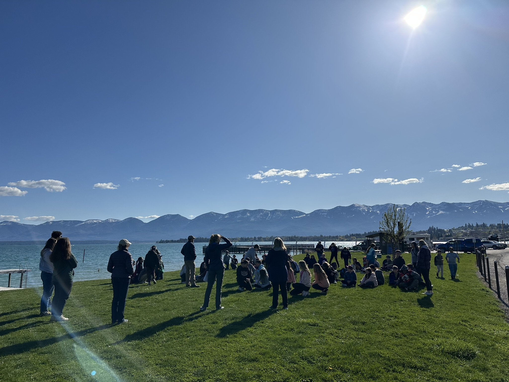 Educators and organization representatives join middle-school students on the shore of Flathead Lake with the Mission Mountains in the background on a sunny day in Salish Point Park, Polson, MT