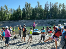 A person is standing on a raft parked on a rocky beach along a river and a crowd of people are around her to learn about pulling noxious weeds for the Weed Rodeo.